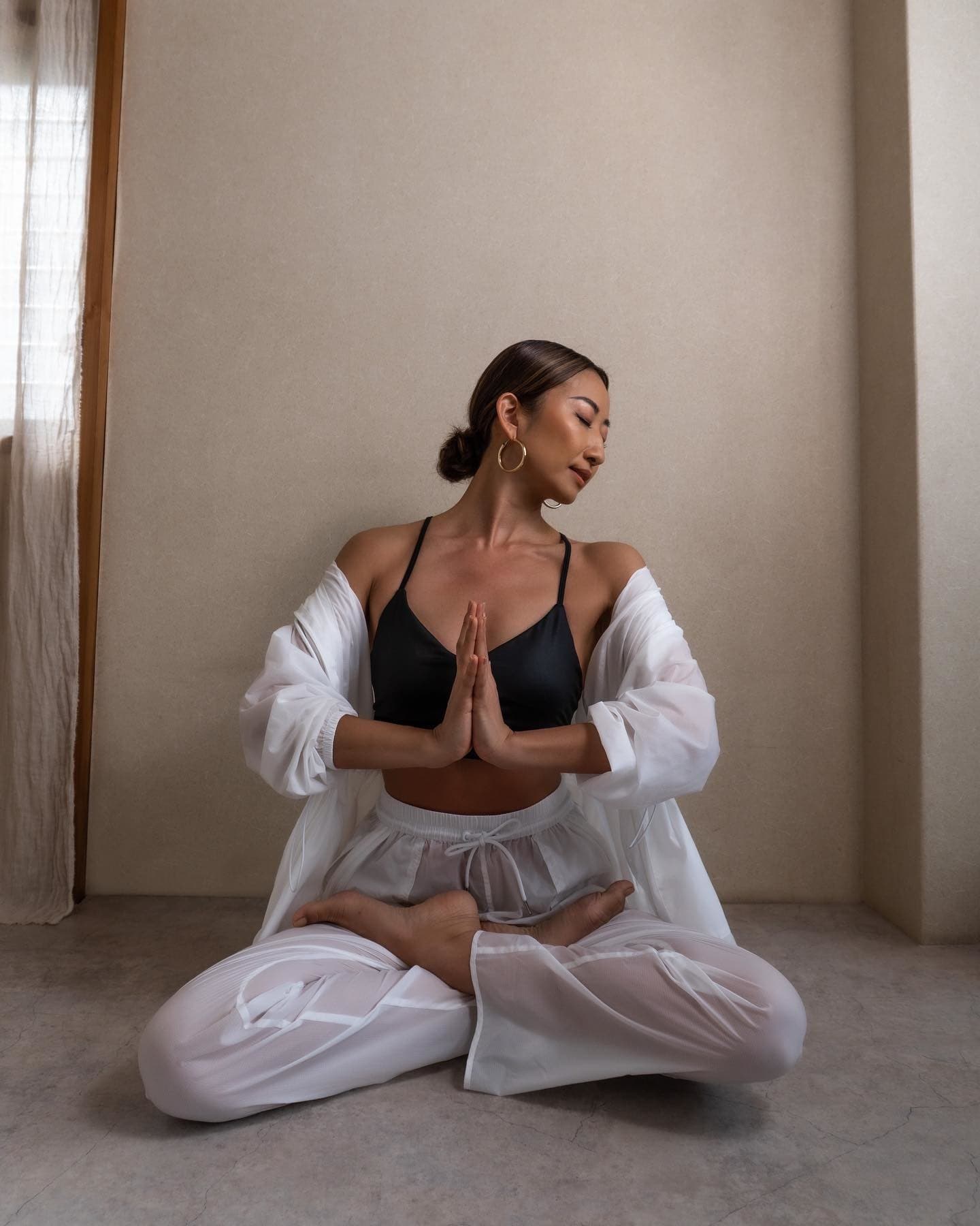@yogibeachhouse wearing a Delight Bralette with a Cloud Nine Jacket and Cloud Nine Pant in white while in Anjali Mudra pose in front of a cream-colored wall.  
