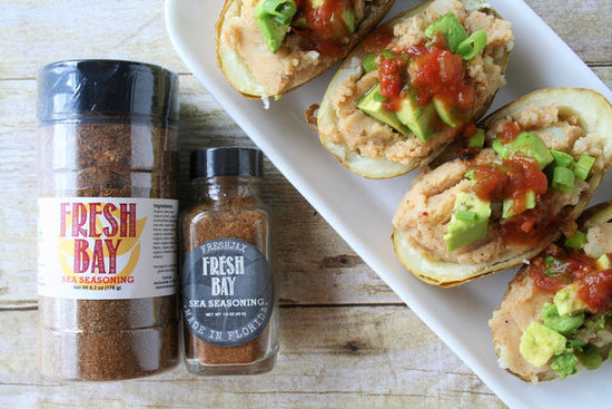 A large and sampler size bottle of FreshJax Organic Fresh Bay Sea Seasoning next to a plate of fresh bay loaded potato skins which are topped with avocado, salsa, and green onions.