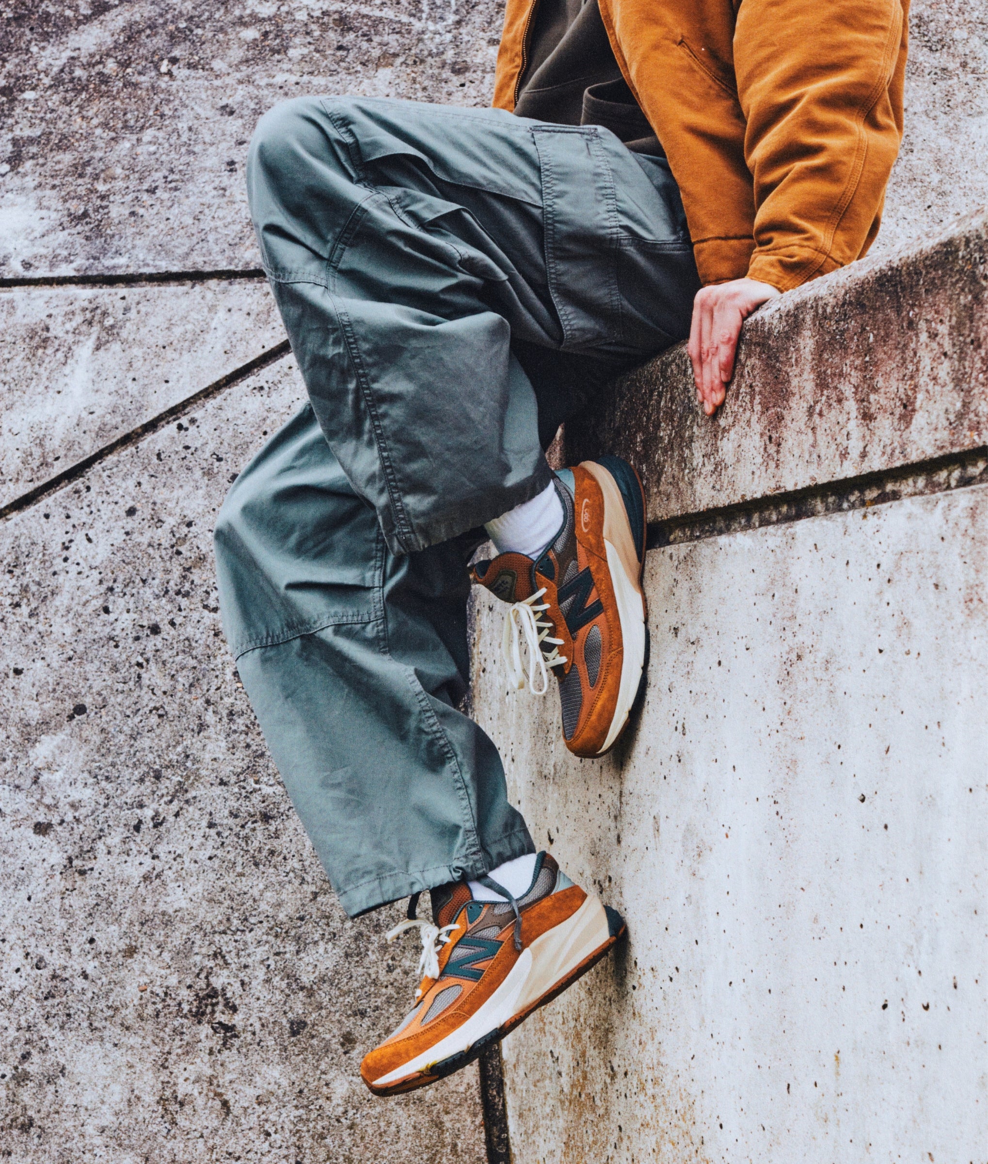 Model sitting on a ledge wearing Carhartt WIP X New Balance 990v6 with Jet Cargo Pants in Smoke Green, and Deep Hamilton Brown OG Detroit Jacket