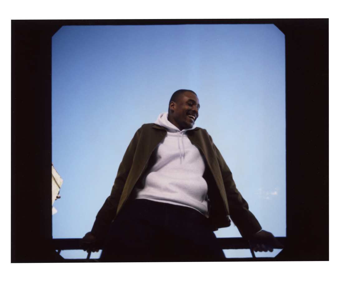 Model with arms holding railing in a white hoodie and brown jacket