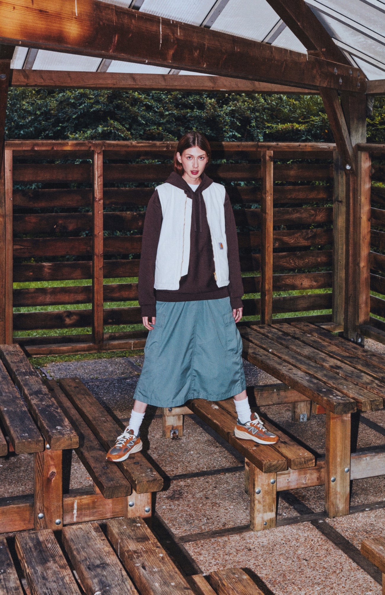 Model standing upon two bench seats wearing Carhartt Vest in Wax, Hoodie in Tamarind, Cargo Skirt in Smoke Green, and 990v6 Carhartt WIP x New Balance Collaboration