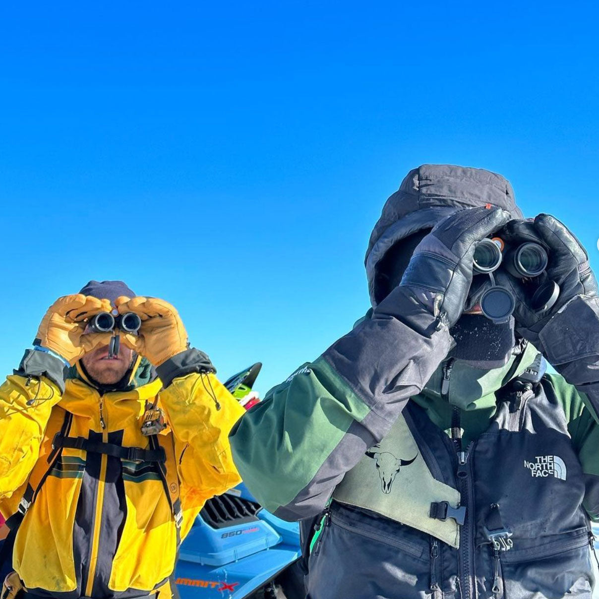 Nils and his friend look up towards the mountain next to their snowmobiles