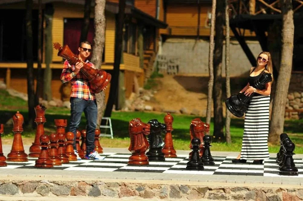Man and woman playing on giant wooden chess set outdoors on black and white tile board surrounded by trees and buildings.