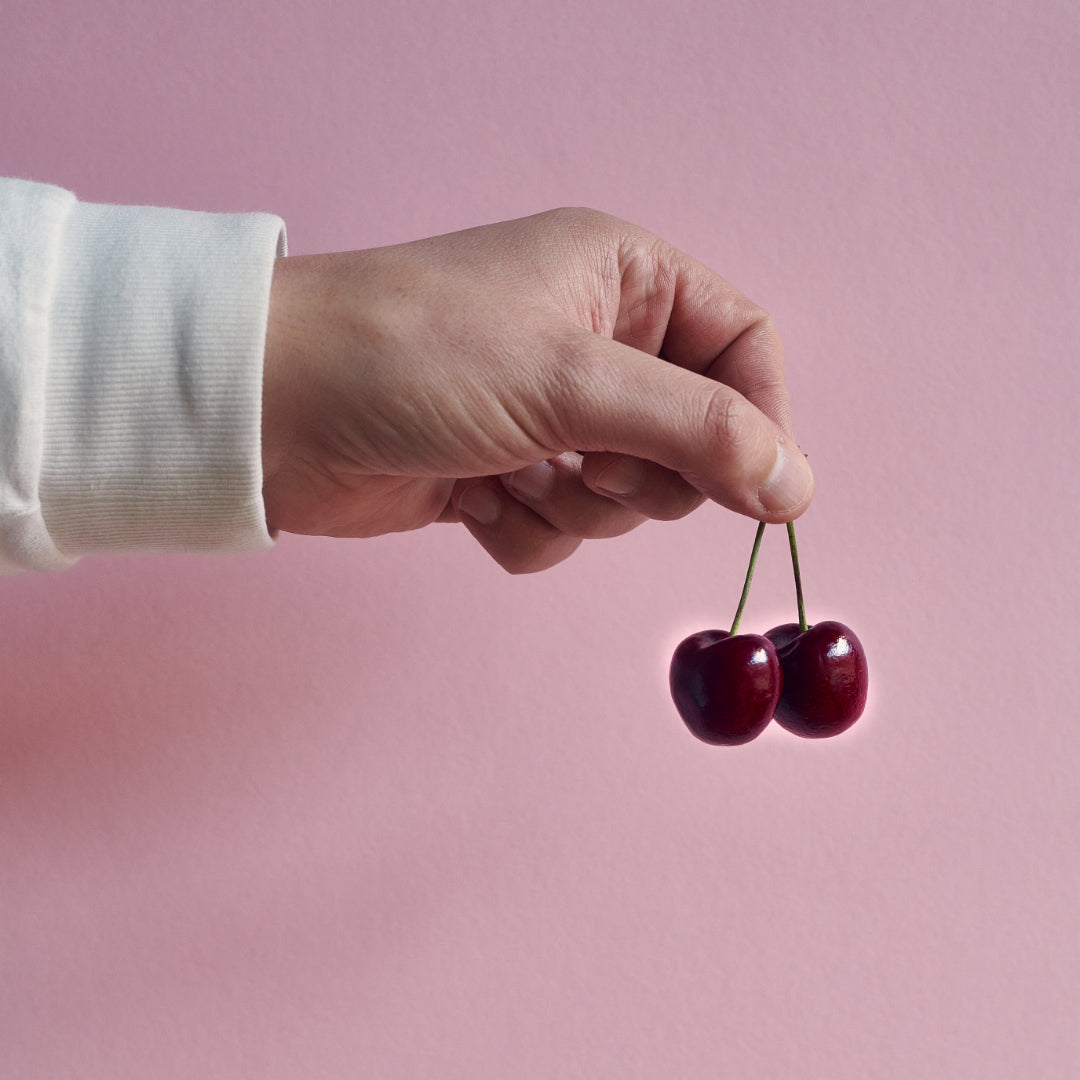 Cherries that are used as an ingredient in Exploding Bakery brownies, being held up against a pink background. 