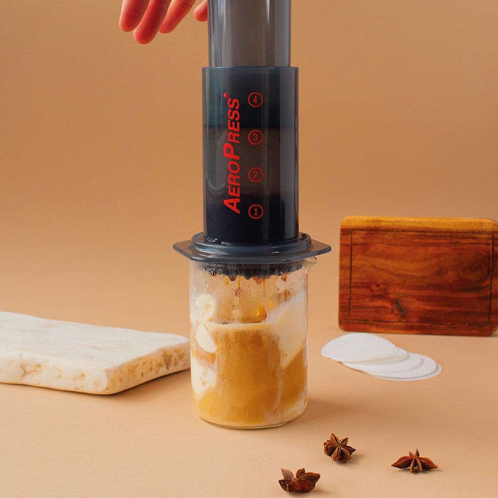 An AeroPress brewing coffee over vanilla ice cream near a granite slab, a serving board, circular filters, and star anise, all on a pecan-colored background.