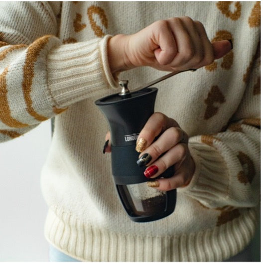 Person using hand coffee grinder to grind coffee beans. Wearing a sweater with print and painted nails
