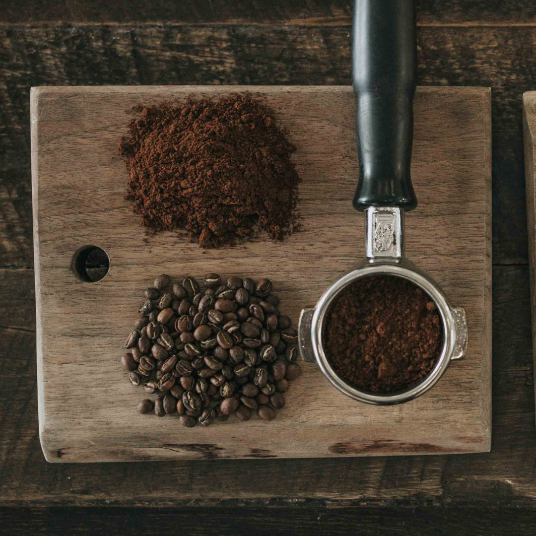 Coffee beans, coffee grounds, and portafilter with coffee grounds on a wooden rustic cutting board on a wooden table