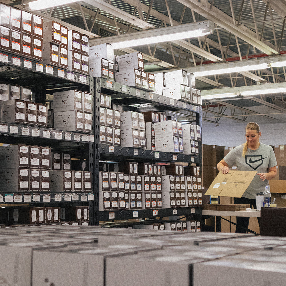 Four-tiered shelves full of coffee pod boxes beside a woman folding a cardboard box.