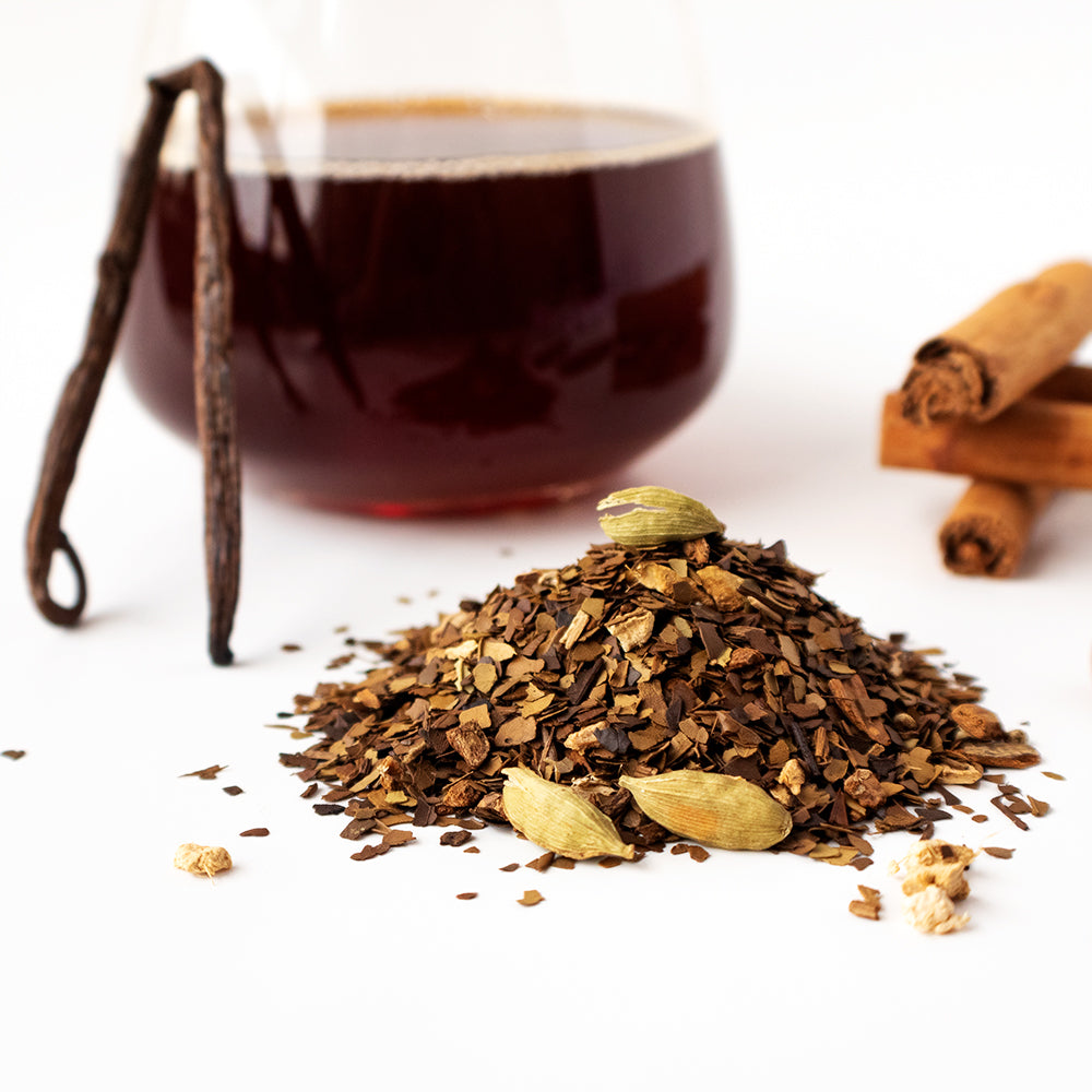 Toasty Mate Chai pile next to a stack of cinnamon sticks, an upright vanilla bean, and a glass of tea.