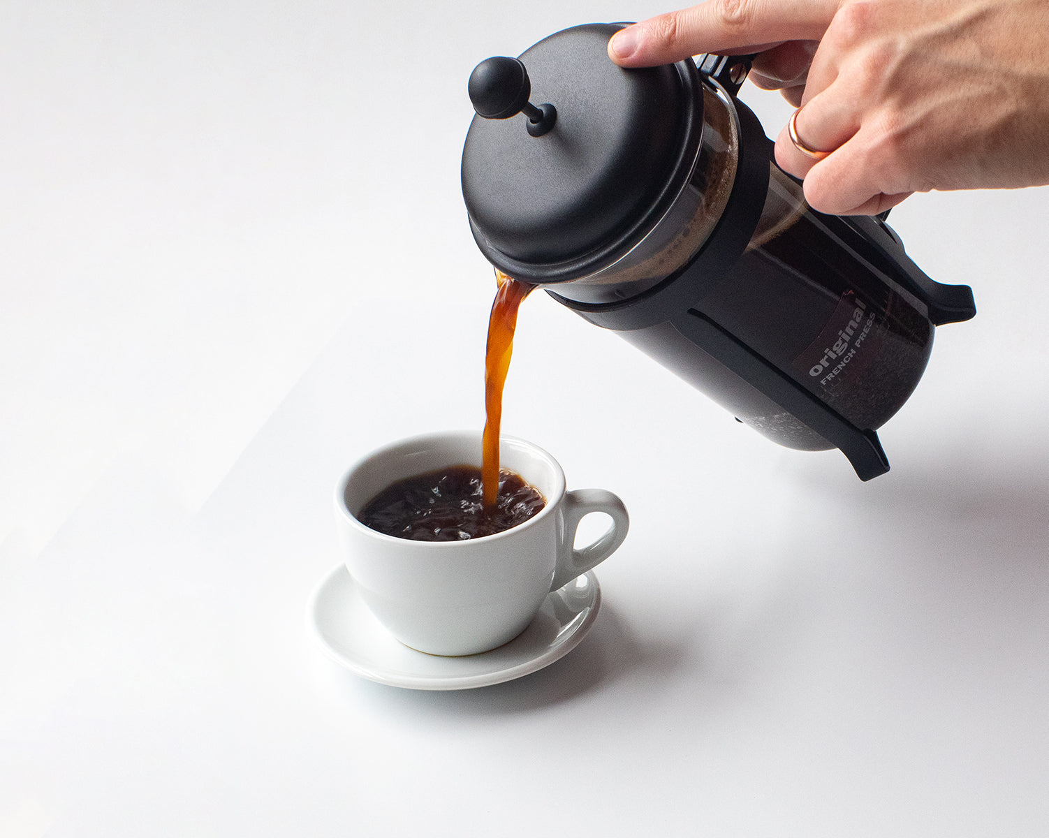A French press pouring coffee into a cappuccino cup on a white background.