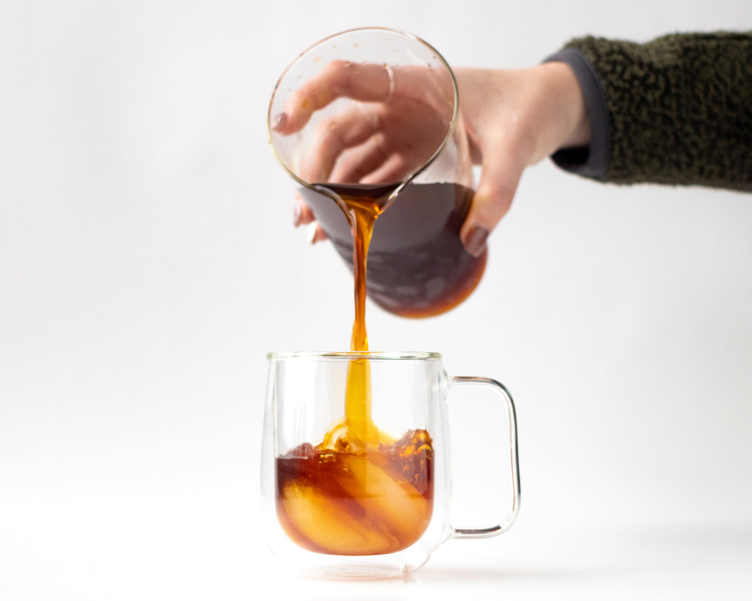 A hand pouring cold brew coffee over ice in a clear mug.