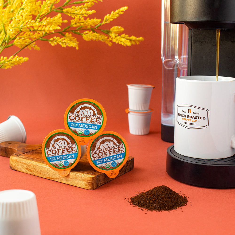 A stack of decaf Mexican pods on a serving board next to a coffee brewer and loose coffee grounds.