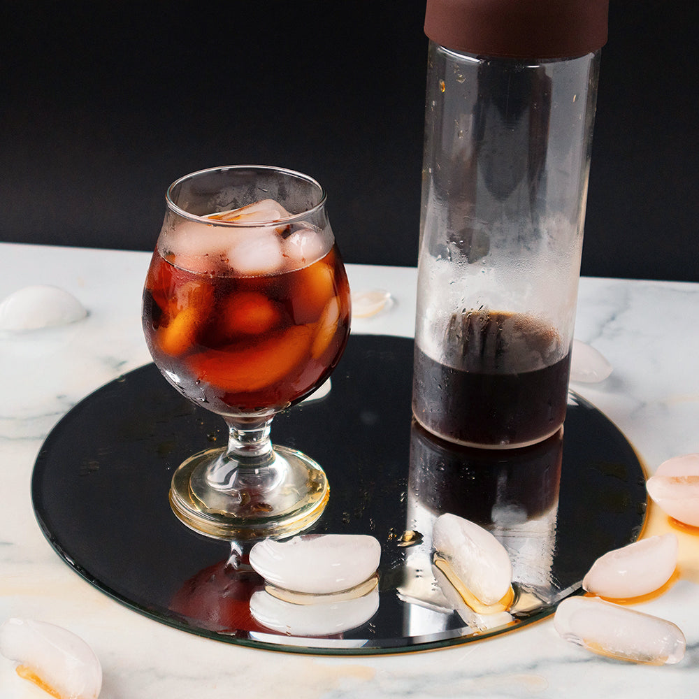 A glass of iced cold brew, a cold brew bottle, and several ice cubes sit on a circular mirror on a granite countertop.