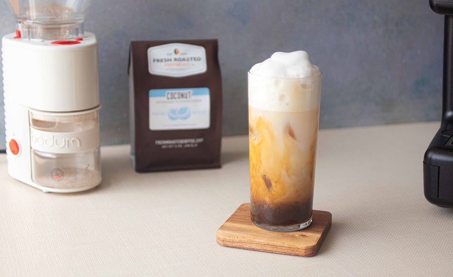 A coconut coffee latte with an espresso machine, bag of coffee, and coffee grinder in the background.