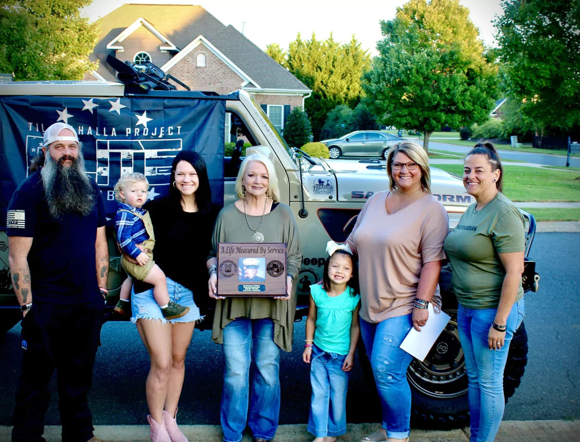 A group of six people and a child posing in front of an off-road vehicle with a military-themed banner.