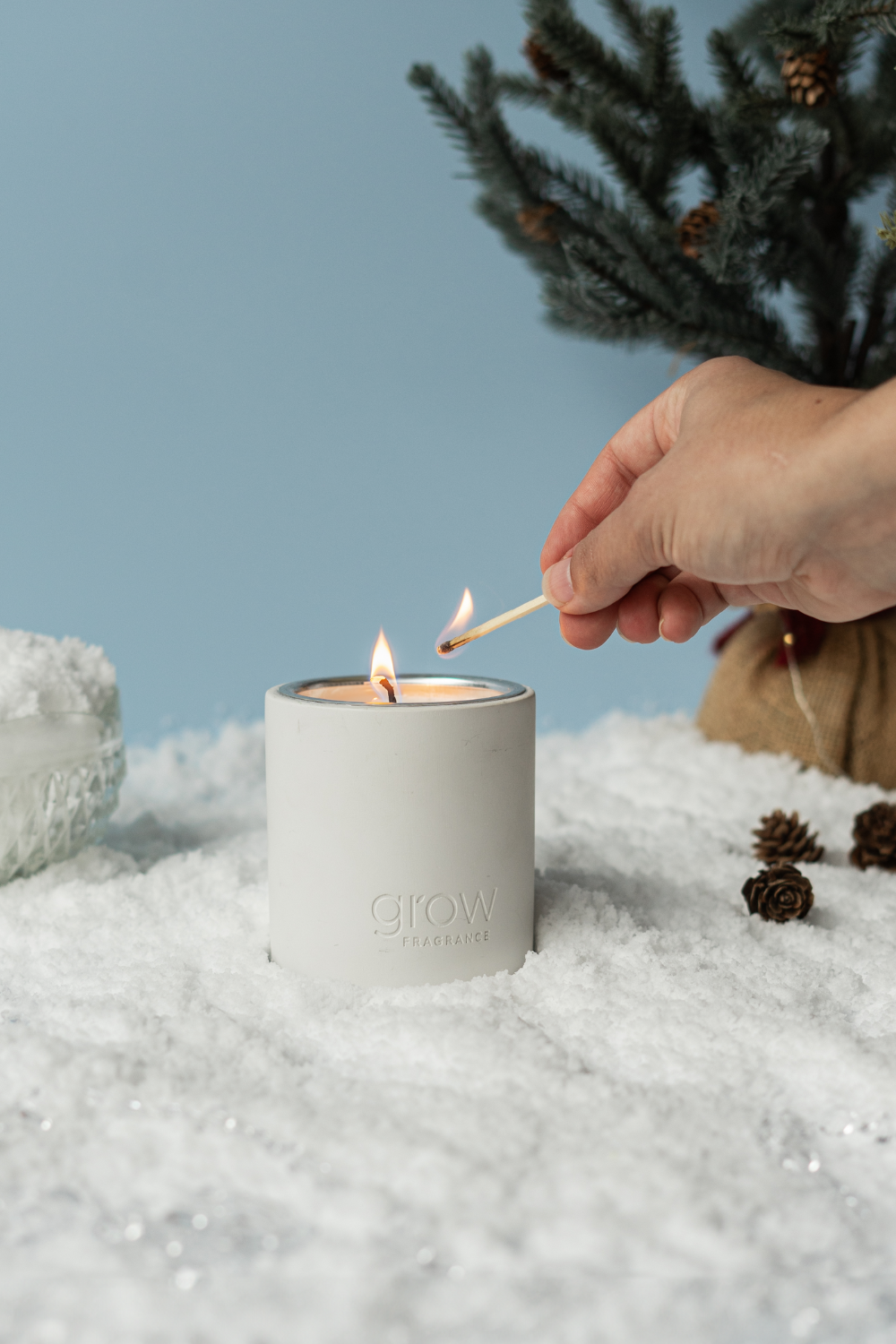 A hand lighting a Snowscape candle against a picturesque winter wonderland backdrop. This candle boasts a delightful blend of vanilla, winter mint, and subtle hints of chocolate, creating a cozy and aromatic ambiance.