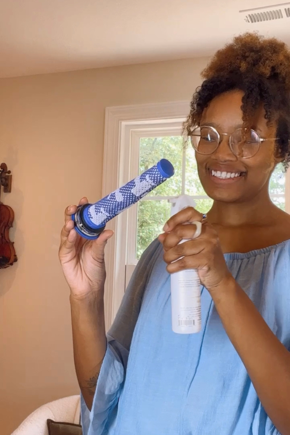 Happy woman in a blue top using a plant-based spray on a blue and white vacuum filter as part of a clever hack to spread holiday scents around the home while vacuuming, standing in a cozy, sunlit room with homey decor.