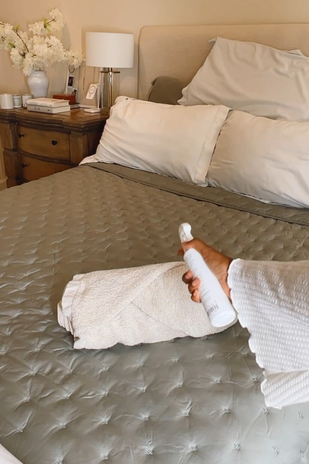 A person's hand holding a white spray bottle and applying a fine mist to a rolled-up towel on a neatly made bed with gray bedding. The bedroom is warmly lit and tastefully decorated with a bedside table featuring a lamp, vase with white flowers, and neatly stacked books, contributing to a serene and inviting atmosphere.