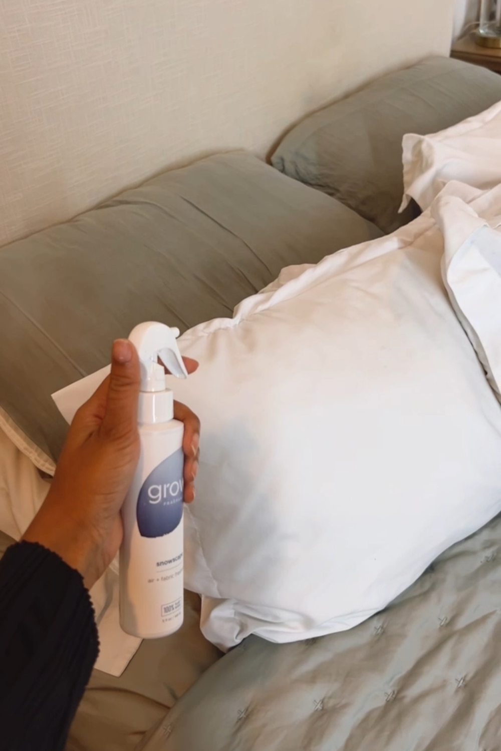 A person's hand is seen holding a white bottle labeled Grow Fragrance Snowscape Scentpoised to spray over a fluffy white pillow. The pillow rests against a backdrop of a neatly made bed with gray sheets and additional plush pillows, creating a cozy and inviting bedroom setting