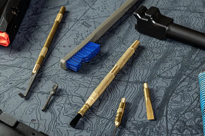 Gun cleaning tools including brass brushes, picks, and scrapers laid out on a topographic-pattern Cerus Gear mat.