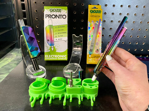 A girl's hand is holding a rainbow Ooze x Stache ConNectar attached to a rainbow Ooze pen. It is aimed into the "e" of the Ooze Stash Station, and a rainbow Ooze Pronto is displayed to the left.