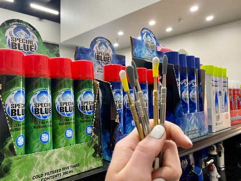 A girl's hand is holding several Ooze dab tools in front of a shelf full of POP displays of cans of butane gas.