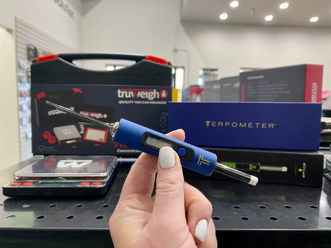 A girl's hand is holding the blue Terpometer thermometer dab tool in front of the Truweigh 710 Pro digital scale kit for concentrates.