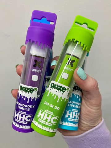 A girl's hand holds a purple, green, and blue package of OozeX HHC disposable 2g vapes. 