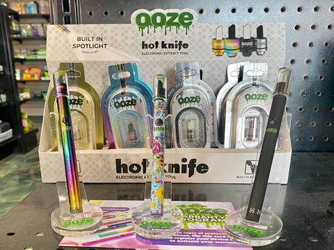A 12ct display of Ooze Hot Knife accessories is displayed behind 3 Twist Slim Pen 2.0 batteries with Hot Knife attachments assembled.