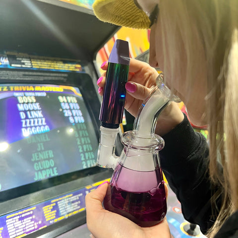 A blonde girl is hitting a purple Glyco freeze bong that has a rainbow Booster inserted as a dab rig.