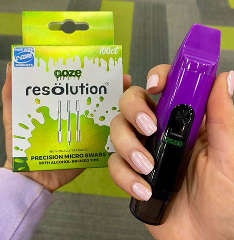 A girl is holding a 100ct box of Ooze Resolution alcohol micro swabs next to a galaxy purple Booster in her other hand.