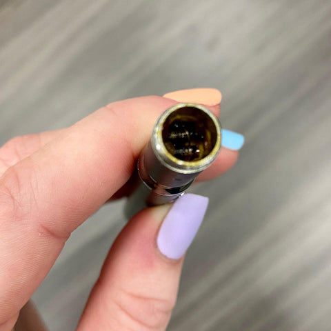 A white female hand with a light purple thumb nail is holding a dirty Ooze wax atomizer coil to show the resin build up on the quartz rods inside.