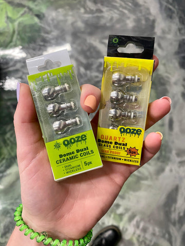 A white female hand is holding two different replacement packs of Ooze domed coils for 510 thread concentrate vaporizers.