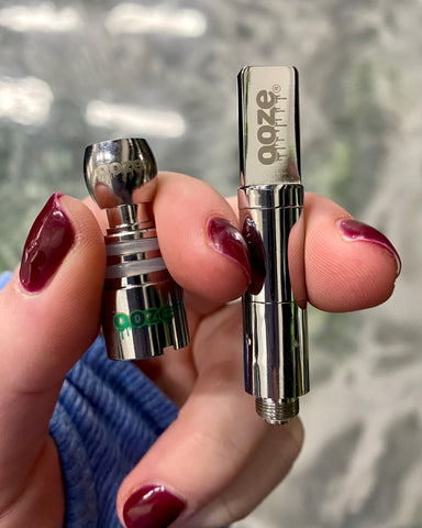 A white female hand with maroon nails holds an Ooze domed coil and an Ooze wax atomizer.