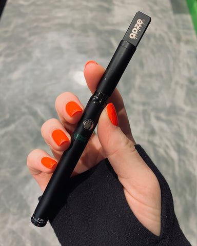 A white female hand with red nails and a black long sleeve shirt with a thumb hole is holding the black Ooze Slim Twist Pro above a gray tile floor. The wax pen has a matching matte atomizer for delta 8 concentrate.