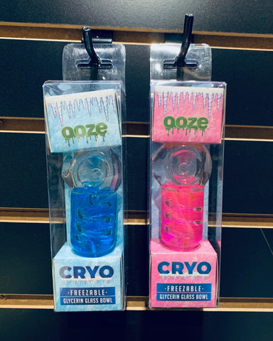 The new blue and pink Ooze Cryo Glycerin Glass bowls are hanging in their full packaging by a hook on a black slat wood wall.