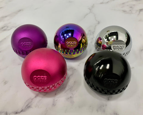 The five new Ooze Saturn globe grinder colors are lined up on a white marble counter. Pink and stealth black are in front with purple, rainbow and chrome behind.