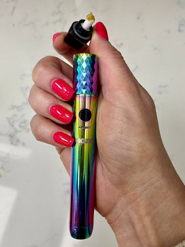 A white girl's hand with bright pink nails is holding a rainbow Ooze Beacon pen with the cap removed and tilted to show a dab of extract on the tool.
