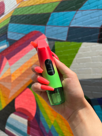 A girl with long red nails holds a Rasta Ooze Booster in front of a colorful mural