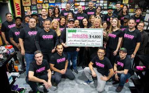 The entire Ooze Wholesale team is gathered in the old showroom and are wearing matching black shirts with pink Ooze logos. In the center is the director from the Shades of Pink Foundation holding a large check for $4,800 which was the Ooze donation.