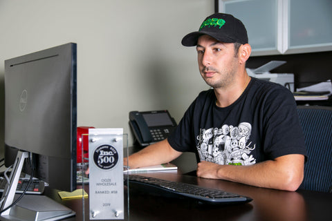 Ooze Wholesale CFO Vince Ayar sits at his desk working on the computer. He wears an Ooze baseball hat, Monsterous shirt, and has the 2019 Inc. 500 plaque just out of focus in the front of the shot.