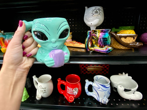 A girl holds up an Alien Mug Pipe in front of a shelf full of other mug pipes