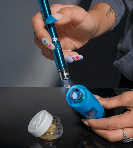 A girl with colorful acrylic nails is using a sapphire blue Ooze Twist Hot Knife to load extract into the atomizer of a blue Ooze Booster. There is an open jar of wax to her left.