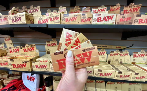A close-up of RAW rolling papers in every size fanned out in a hand that is in front of a whole wall full of RAW products.