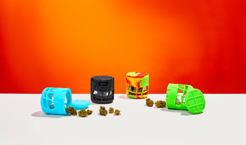 4 silicone and glass jars are all filled with weed and lined up together on a white table. The teal and green are on the ends, with the lids off and spilling over. The black and Rasta are centered with the lids on.