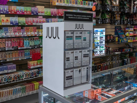 A Juul POP display is sitting on a glass counter in front of a wall display of other disposable nicotine vapes.