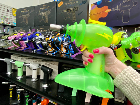 A girl holds the green Thicket Spaceout Ray Gun butane novelty torch in front of a shelf of torches in the Cannatron wholesale showroom