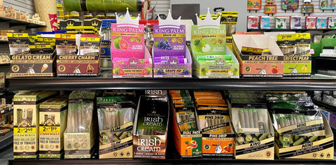 An assortment of King Palm pre-rolled cone POP displays are lined up on two black shelves in a smoke shop.