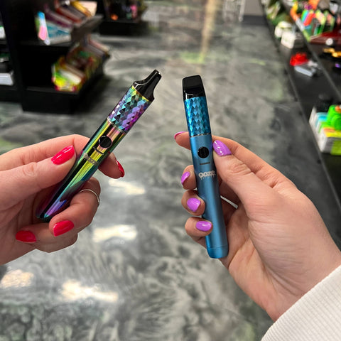 Two girls are holding their Ooze Beacon extract vapes together in the Cannatron showroom. The Beacon on the right is blue and the left one is rainbow.