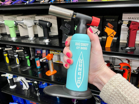 A girl holds a light teal Blazer Big Shot torch in front of a shelf of torches in the Cannatron wholesale showroom